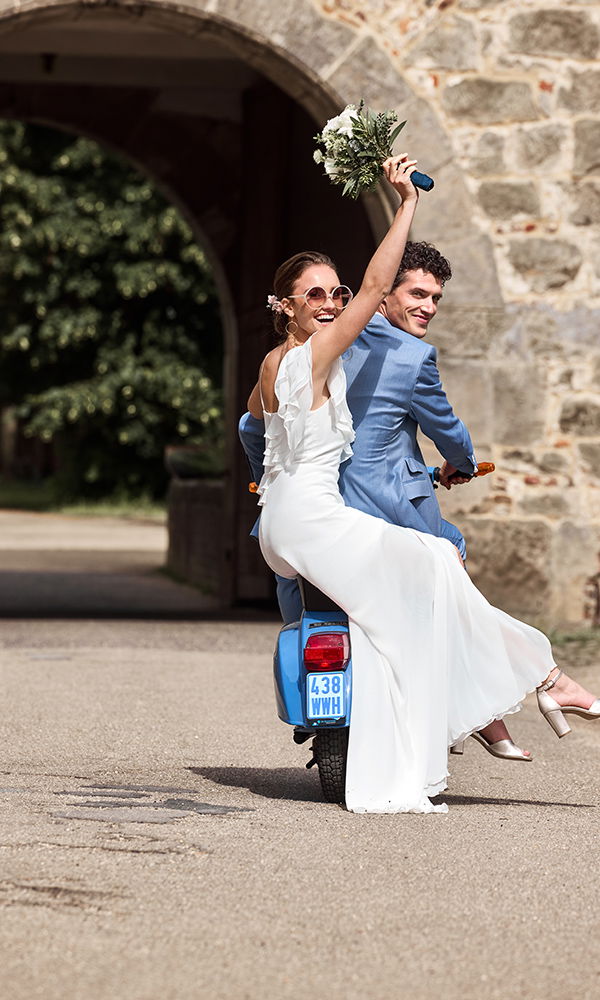 Frisch verheiratetes Paar fährt fröhlich auf einem blauen Roller davon, die Braut hält den Brautstrauß in die Luft.