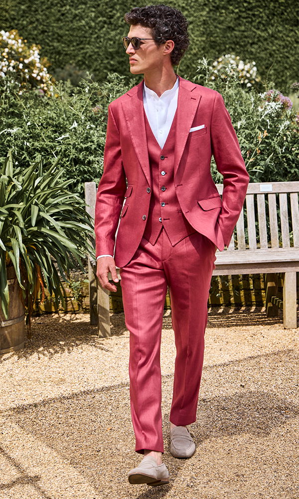 Mann in einem eleganten, roten Anzug mit Weste und weißem Hemd, schlendert stilvoll durch einen Garten.