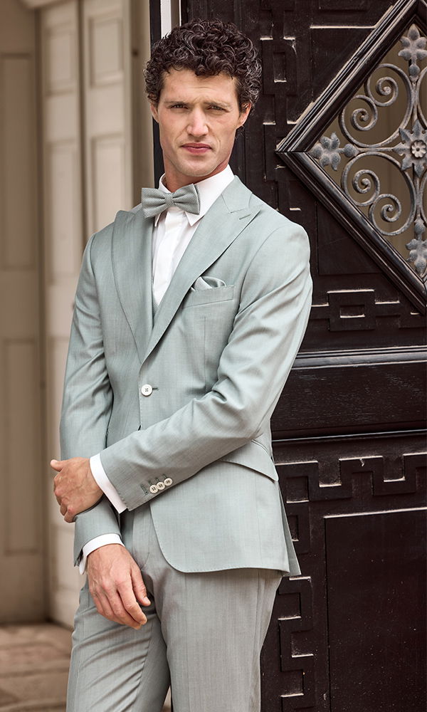 Mann in einem salbeigrünen Anzug mit Fliege und weißem Hemd posiert vor einem verzierten, dunklen Holztor.