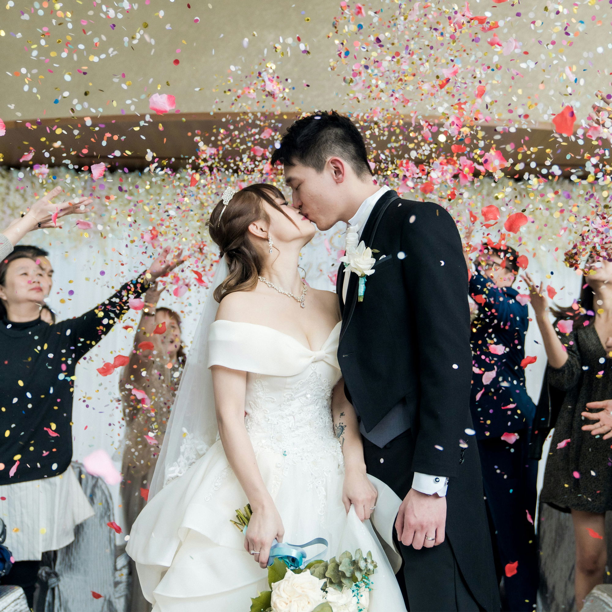 Hochzeitsszene mit küssendem Brautpaar in eleganter Kleidung und Konfettiregen im Hintergrund, Gäste feiern mit.
