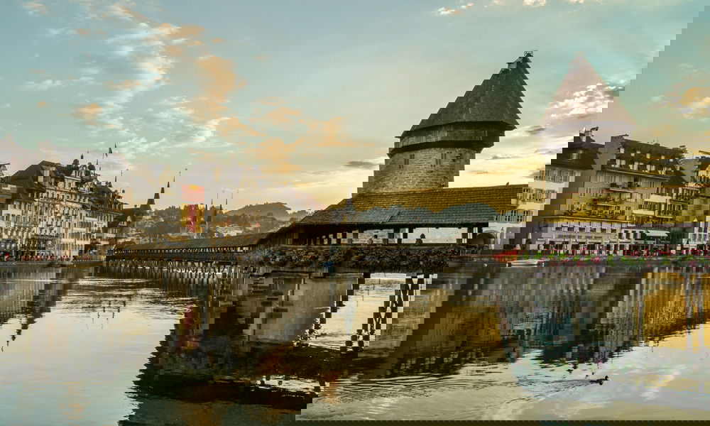 Die historische Kapellbrücke mit Wasserturm in Luzern bei Sonnenuntergang, gespiegelt im ruhigen Wasser der Reuss.