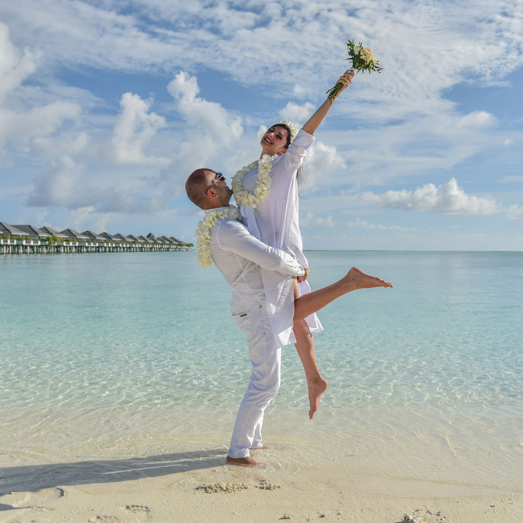 Verliebtes Paar in Weiß posiert am türkisblauen Wasser mit leisem Urlaubsflair und tropischer Kulisse.