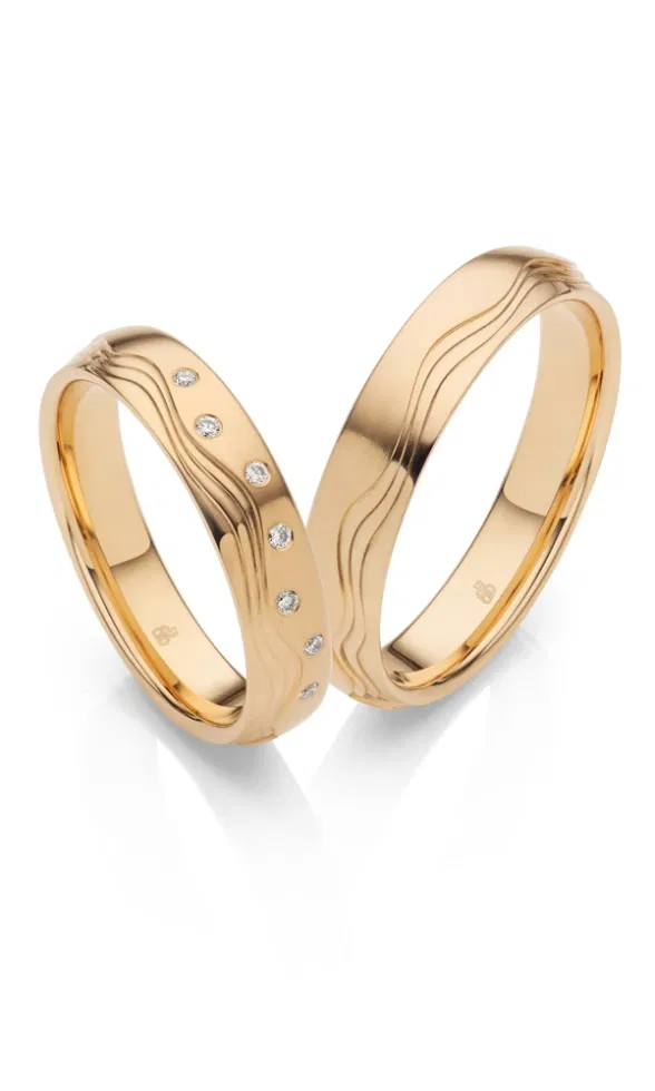 Zwei goldene Trauringe mit matter Oberfläche und sanfter Wellenstruktur, ein Ring zusätzlich mit mehreren dezent gefassten Diamanten verziert.