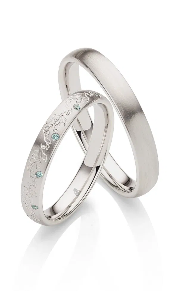 Zwei silberfarbene Trauringe mit matter Oberfläche, einer schlicht, der andere mit filigraner Blättergravur und mehreren dezent schimmernden Edelsteinen verziert.