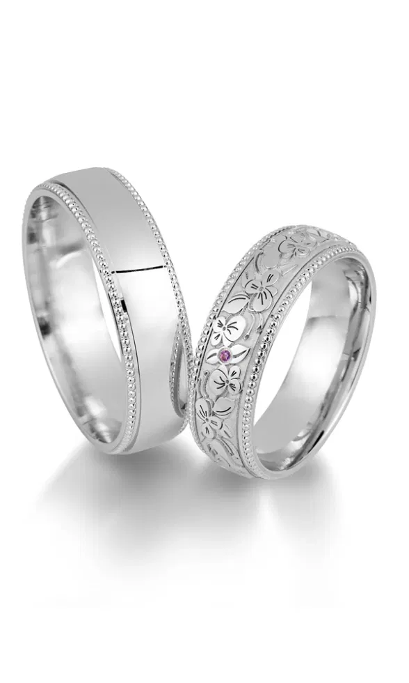 Zwei silberfarbene Trauringe mit glänzender Oberfläche und feiner Perlenkante; ein Ring ist mit floraler Gravur und einem kleinen farbigen Edelstein verziert.