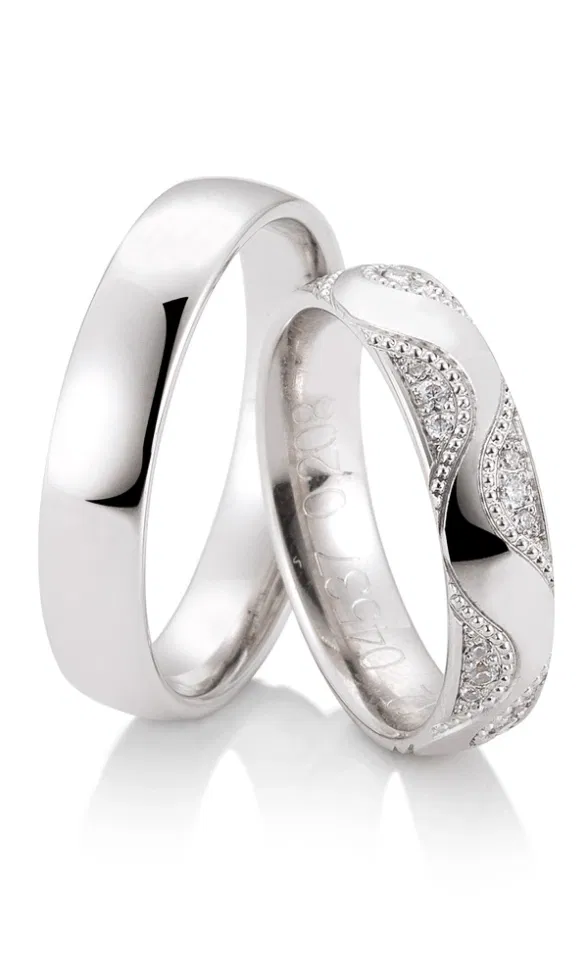 Klassische Trauringe aus hellem Edelmetall, ein Ring schlicht poliert, der andere mit geschwungenem Muster und funkelndem Diamantbesatz.