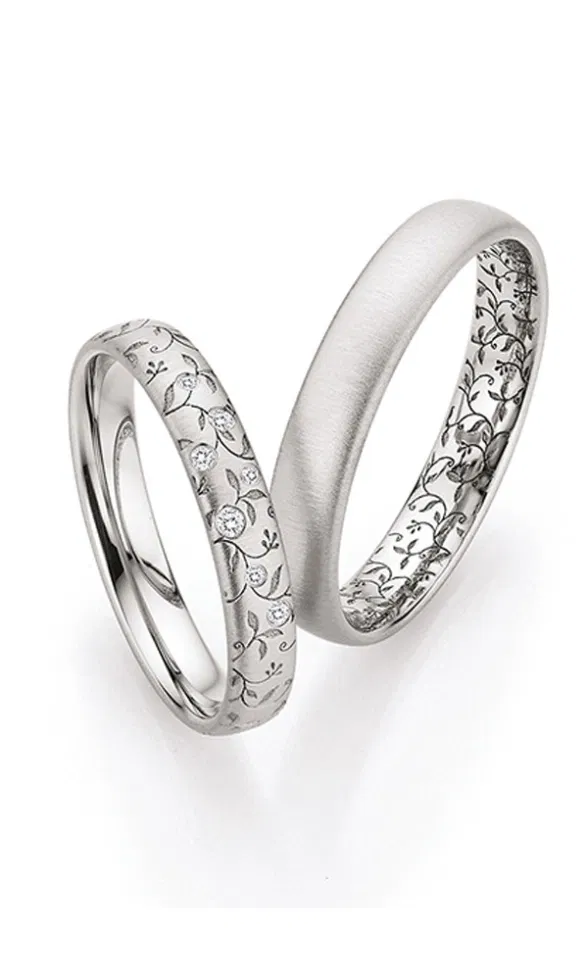 Zwei silberfarbene Trauringe mit matter Oberfläche, feiner floraler Gravur und mehreren kleinen Diamanten, die das romantische Design unterstreichen.