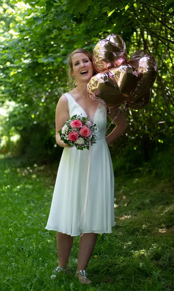Braut in Chiffonkleid posiert mit Herzballons und Blumenstrauß unter schattigen Bäumen.