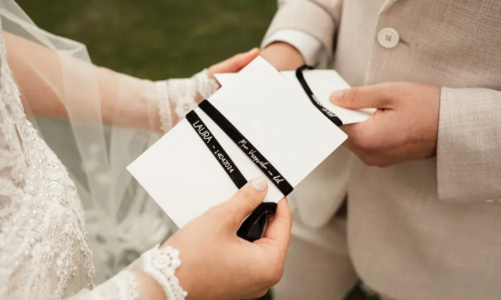 Ein Brautpaar hält jeweils eine Klappkarte mit schwarzer Banderole in den Händen und liest sich ihr Eheversprechen gegenseitig vor.