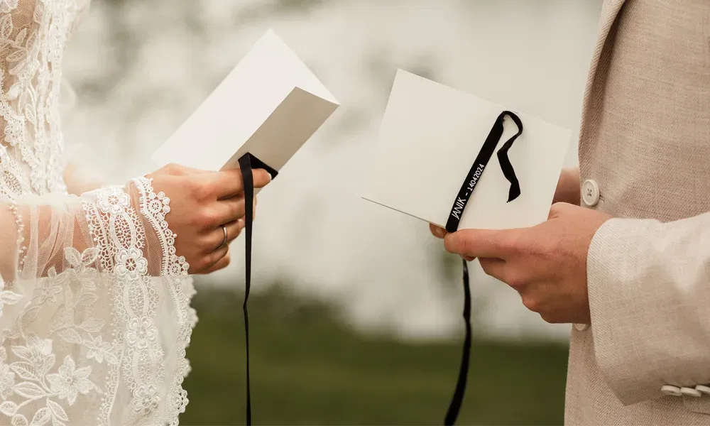 Ein Brautpaar hält jeweils eine Klappkarte mit schwarzer Banderole in den Händen und liest sich ihr Eheversprechen gegenseitig vor. Sie stehen sich gegenüber.