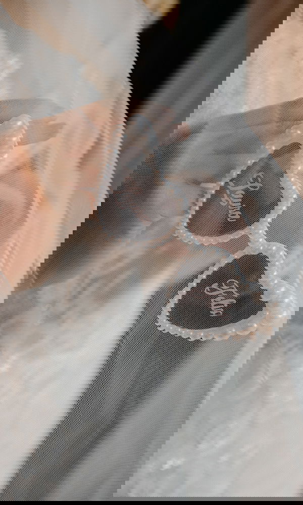 Stylisches Hochzeitsdetail: Herzbrille mit Perlenbesatz und Namen, gehalten unter einem Brautschleier.