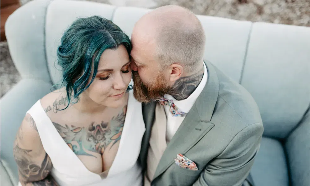 Das Brautpaar sitzt vertraut auf einem Sofa – er küsst sie sanft an der Schläfe, sie wirkt entspannt mit geschlossenen Augen. Auffällig sind ihre Tattoos, sein florales Einstecktuch und ihre intensiven Haarfarben.