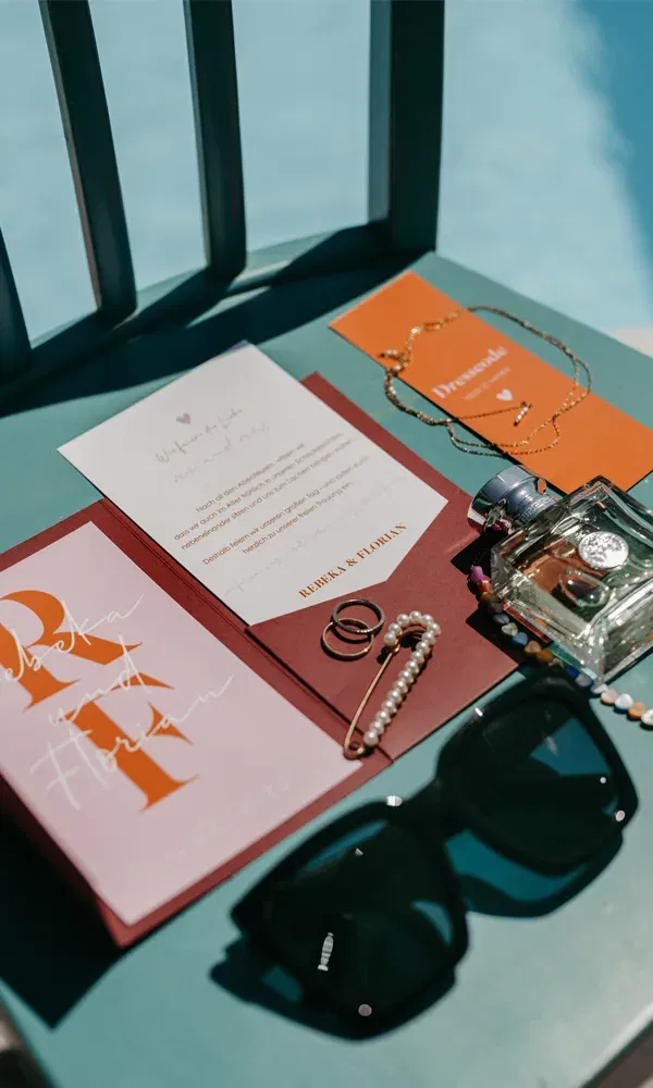 Einladung zu Rebeka & Florians Fest mit Accessoires wie Ringen, Schmuck und Sonnenbrille auf einem türkisfarbenen Tisch.