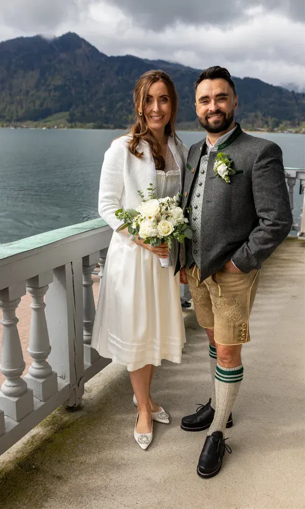 Janine und Martin an ihrem Hochzeitstag am Tegernsee, sie im weißen Dirndl mit Blumenstrauß, er in Tracht mit Hirschlederhose.