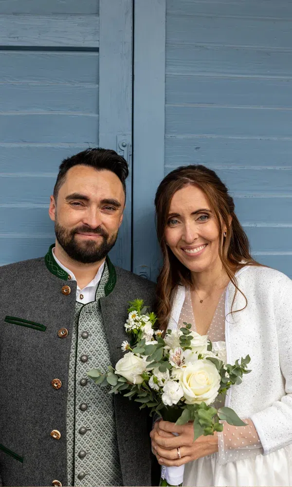 Janine und Martin bei ihrer Trauung am Tegernsee – sie mit Blumenstrauß in Weiß, er in bestickter Lederhose und Janker.