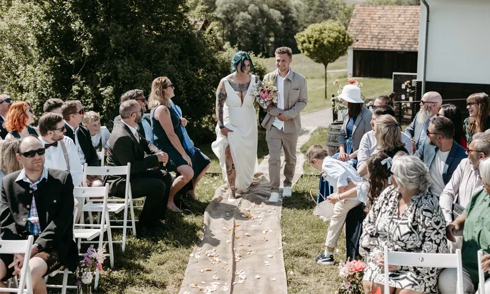 Rebeka wird bei der Trauung von ihrem Sohn begleitet, während sie den Gang entlanggeht – herzliche Blicke und Sommerstimmung im Freien.