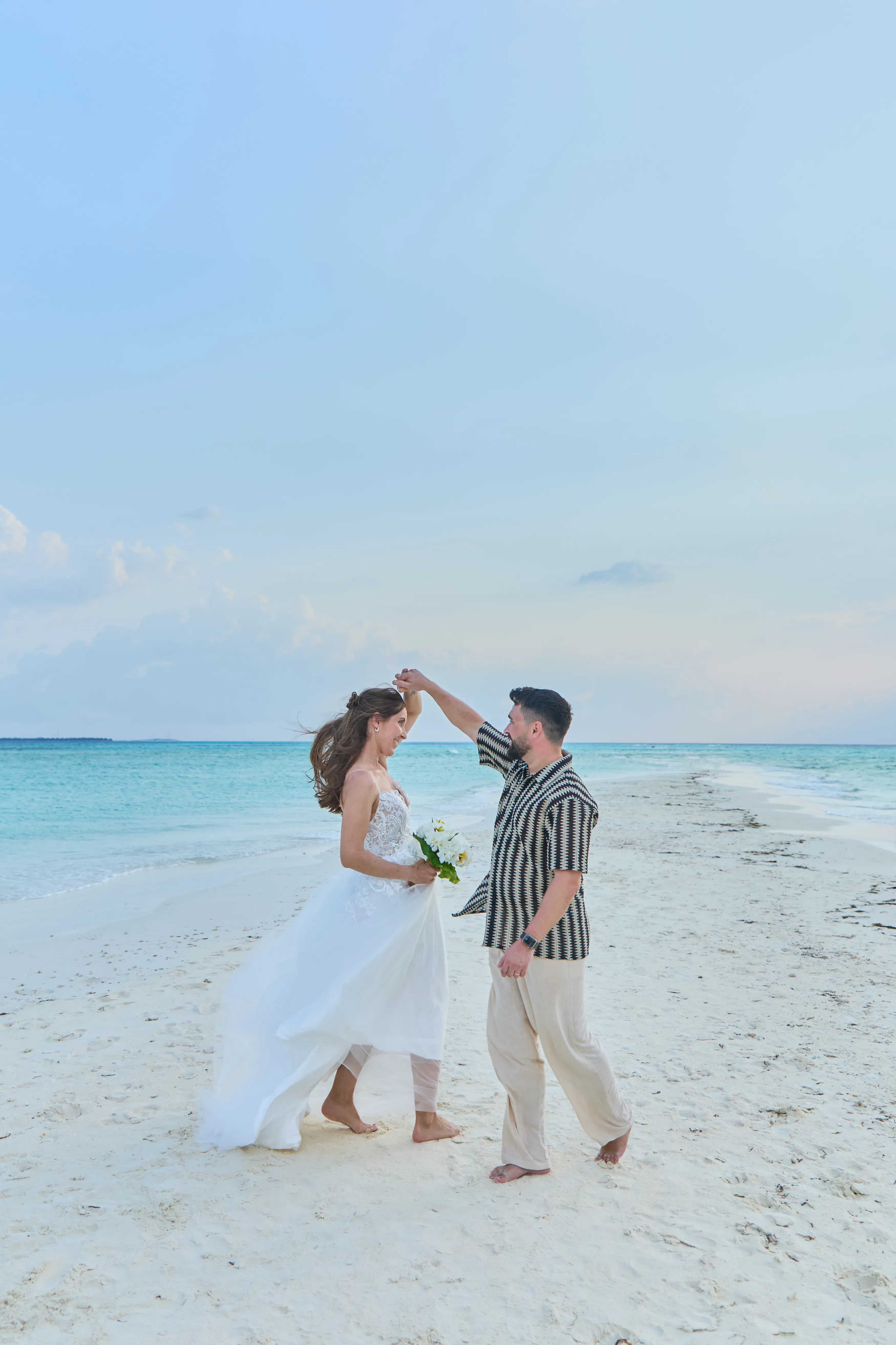 Brautpaar beim Tanzen am Strand – luftiges Hochzeitskleid, barfuß und voller Freude auf einer schmalen Sandbank im Paradies.