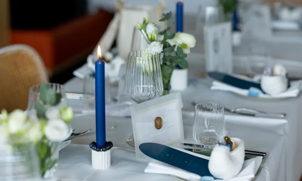 Tischdeko in Blau-Weiß bei einer Hochzeitsfeier: brennende Kerze, Blumenschmuck, Menükarte mit Siegel und Gläser auf weißer Tischdecke.