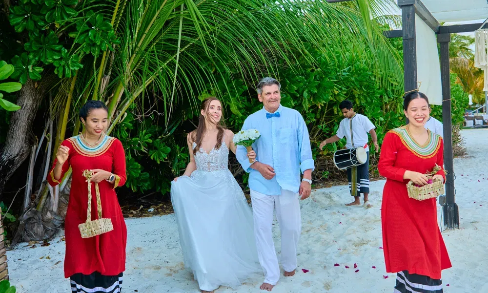 Hochzeitszug am Strand: Die Braut wird von ihrem Vater geführt, mit Blumen, Musik und festlicher Atmosphäre unter Palmen.