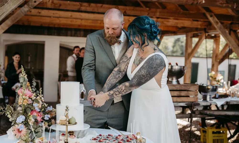 Gemeinsames Anschneiden der Torte beim Wedding-Weekend: Braut mit blauen Haaren und tätowierten Armen, Bräutigam im grauen Anzug.