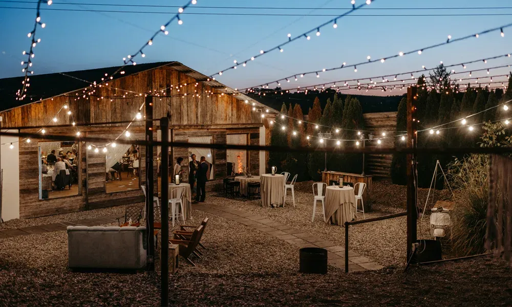 Gemütliche Scheunenhochzeit bei Sonnenuntergang: Lichterketten und festlich gedeckte Tische sorgen für stimmungsvolles Ambiente.