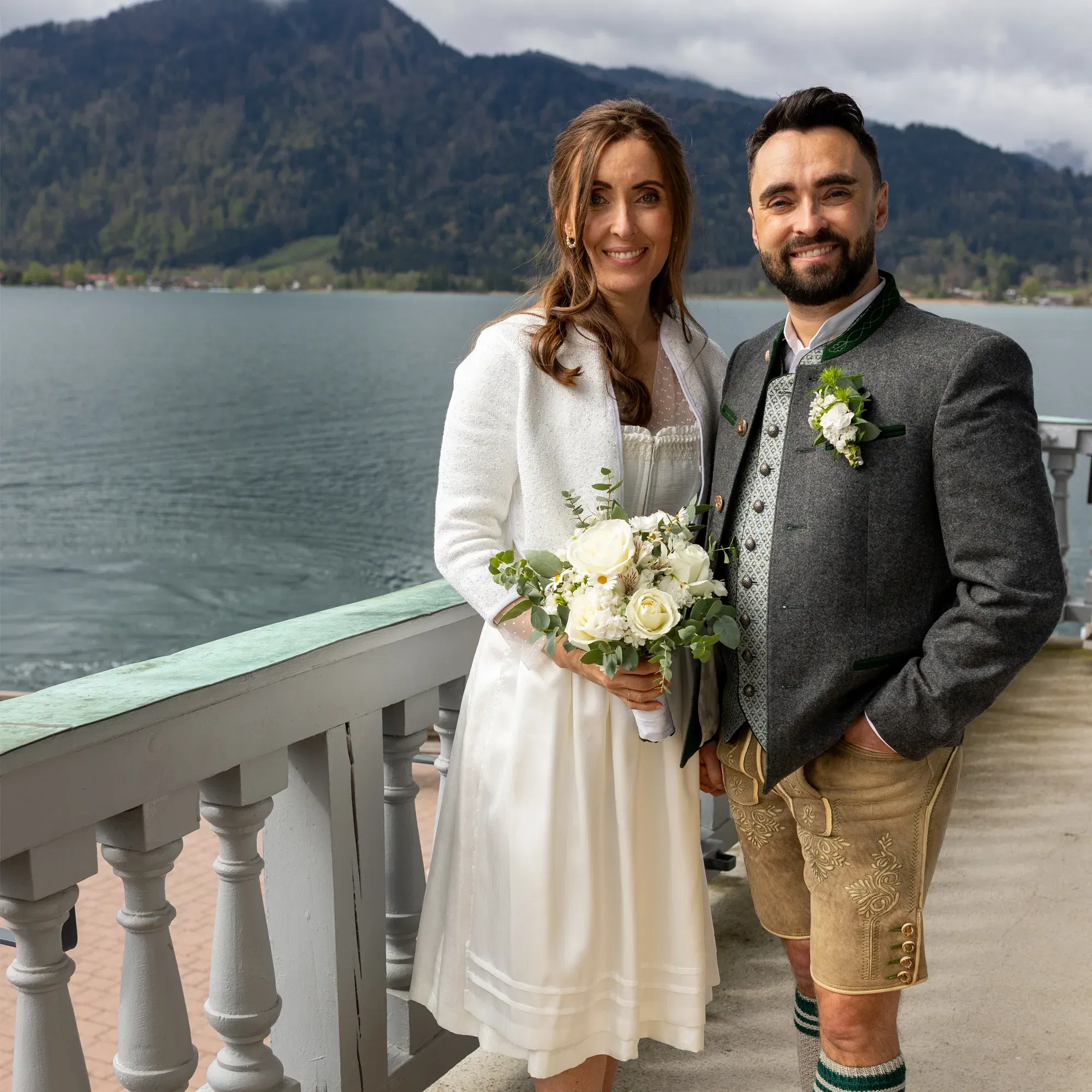 Janine und Martin an ihrem Hochzeitstag am Tegernsee, sie im weißen Dirndl mit Blumenstrauß, er in Tracht mit Hirschlederhose.