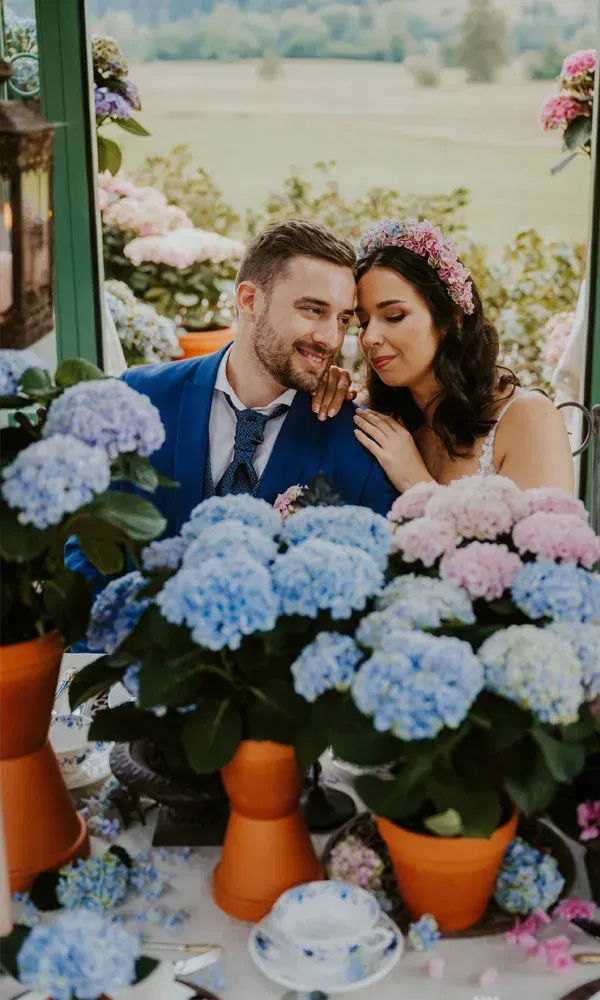 Zwischen blauen und rosafarbenen Blüten: Ein verliebtes Brautpaar an einem blumengeschmückten Hochzeitstisch.