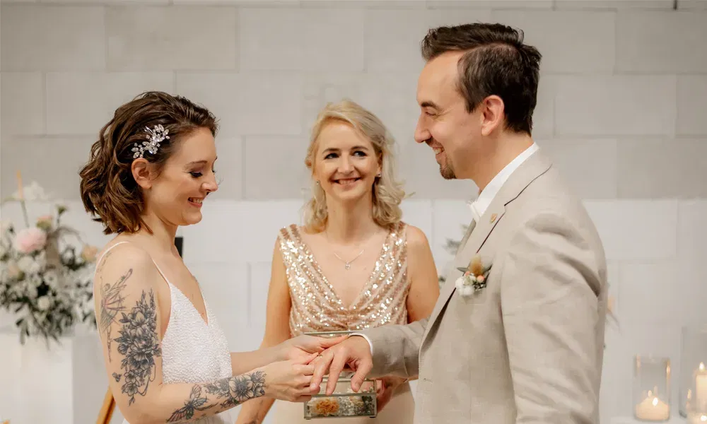 Emotionale Ringtausch-Szene bei einer Hochzeit: Die Rednerin im glitzernden Kleid begleitet das Brautpaar mit liebevollem Blick.