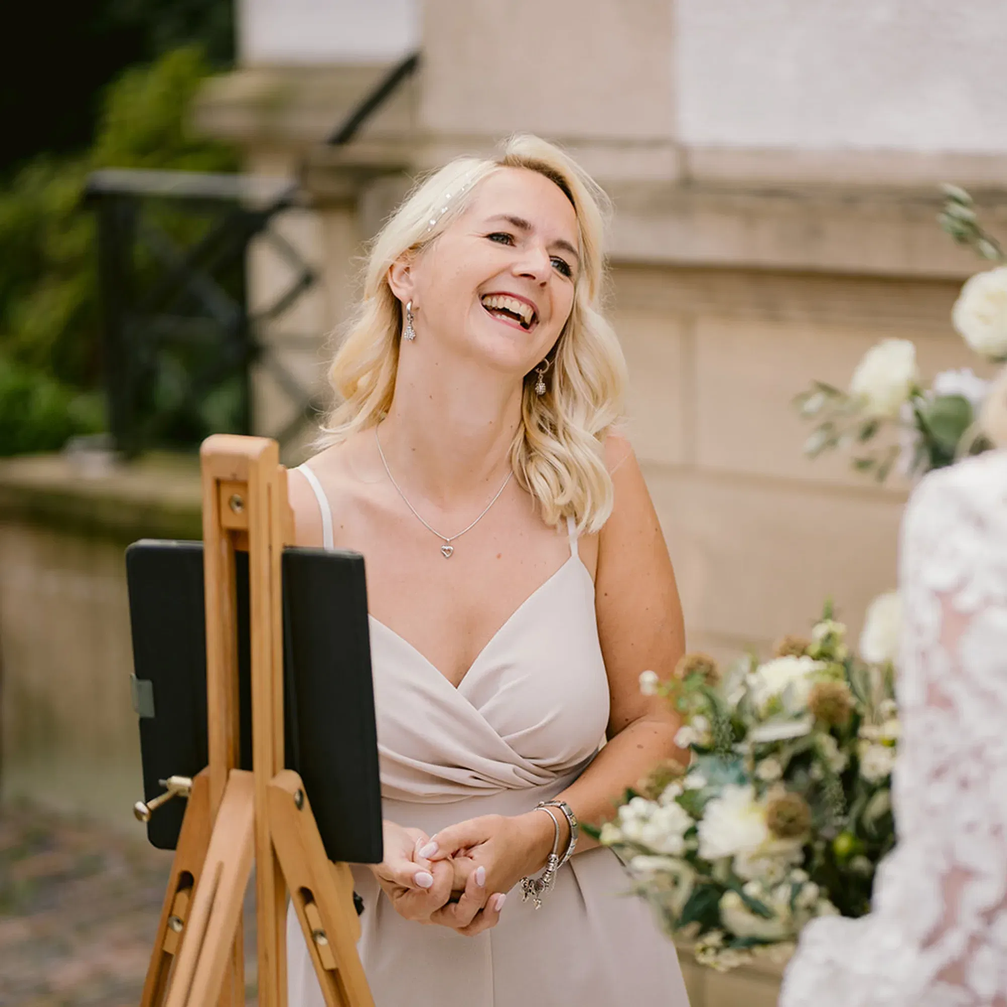 Emotionale Szene bei einer freien Trauung: Die Rednerin lacht strahlend, Braut mit Blumen im Vordergrund.