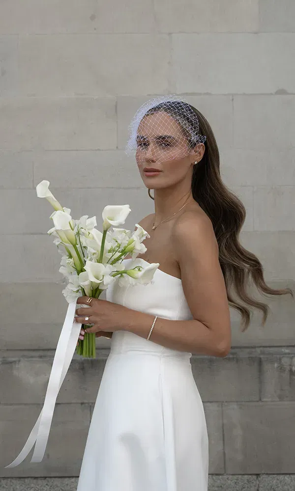 Braut mit langen Haaren und Schleier im Netzdesign hält ihren weißen Calla-Strauß vor heller Steinwand.