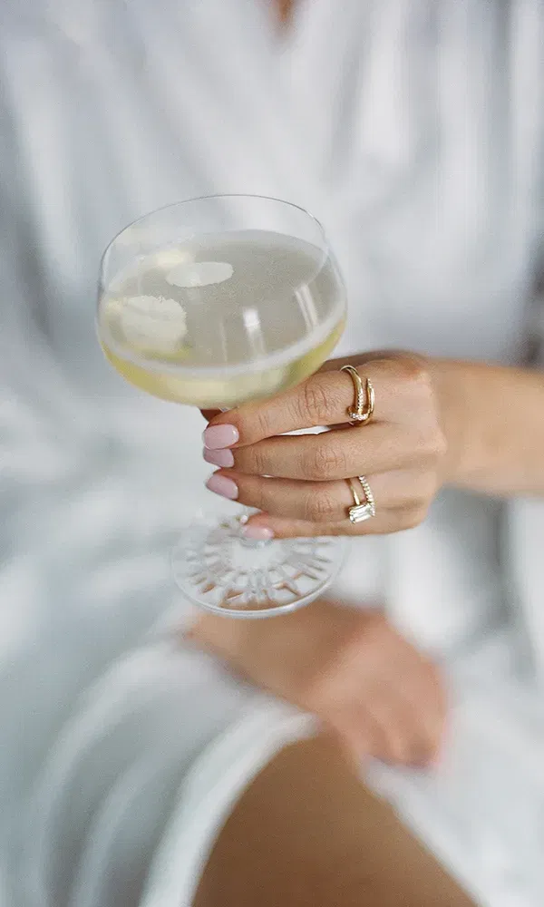 Hand mit Eheringen und zartem Nageldesign hält ein elegantes Sektglas – stilvoller Hochzeitsmoment.