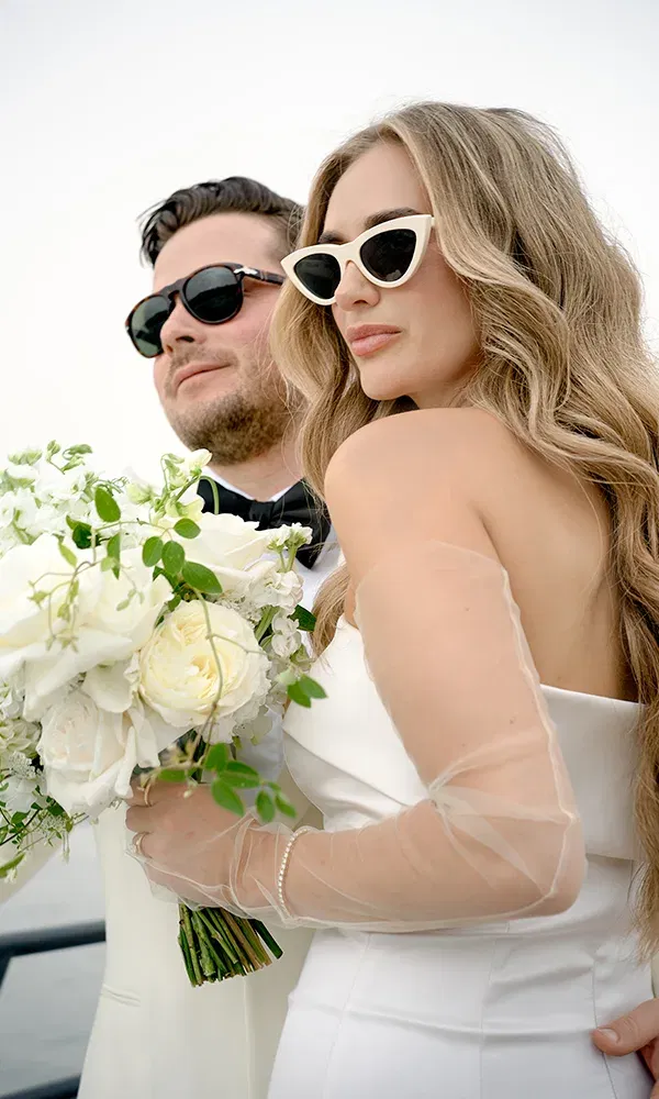 Stylishes Brautpaar mit Sonnenbrillen posiert auf einem Boot mit weißem Blumenstrauß – modern und glamourös.
