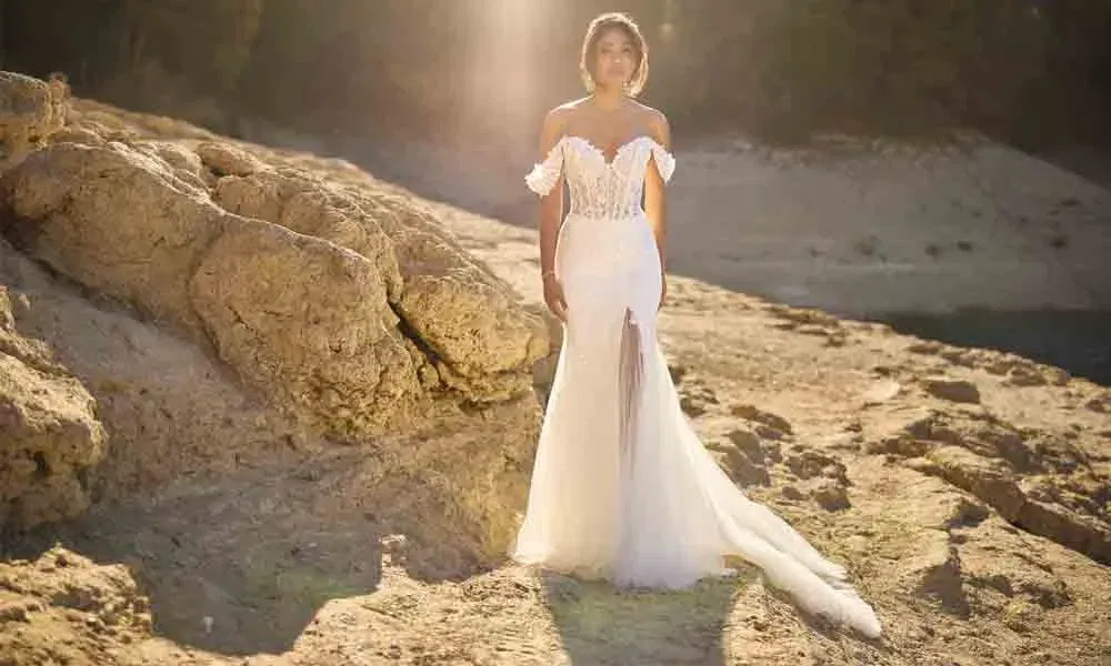 Braut in figurbetontem Kleid mit transparenter Korsage und fließender Schleppe, fotografiert in der Abendsonne auf rauem Gestein.