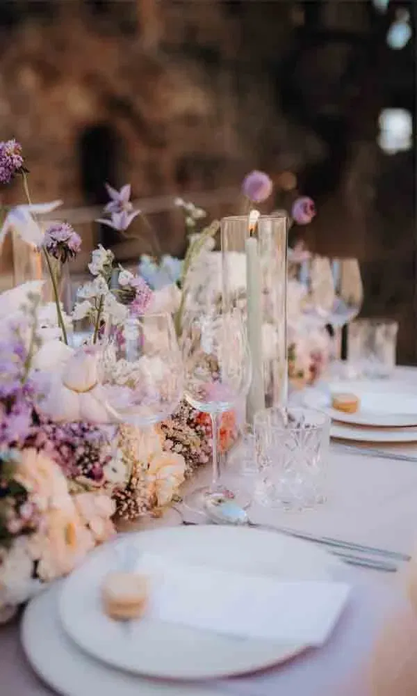 Detailaufnahme einer eleganten Hochzeitstafel mit Glasdekoration, Kerzen und pastellfarbenen Blumen in Violett- und Rosatönen.