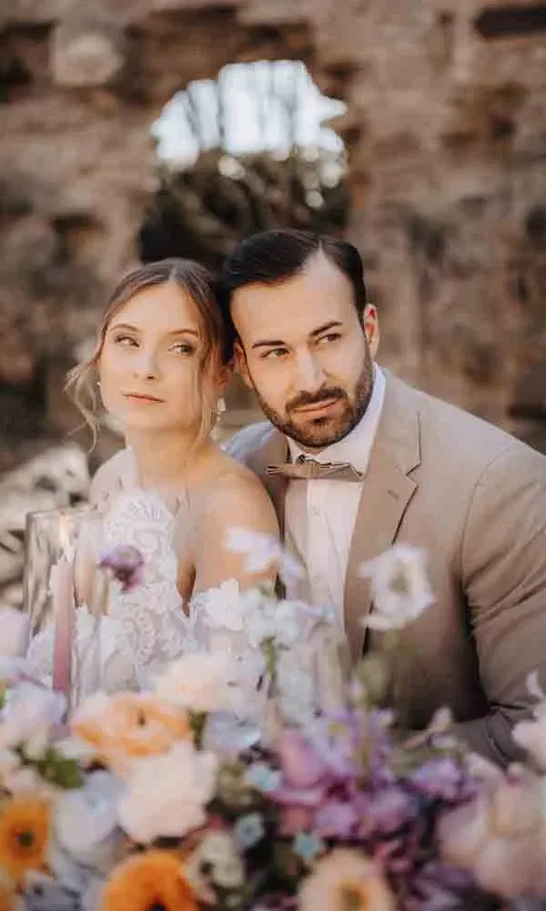 Braut und Bräutigam sitzen an einem festlich geschmückten Tisch, umgeben von bunter Frühlingsfloristik vor rustikalem Mauerwerk.