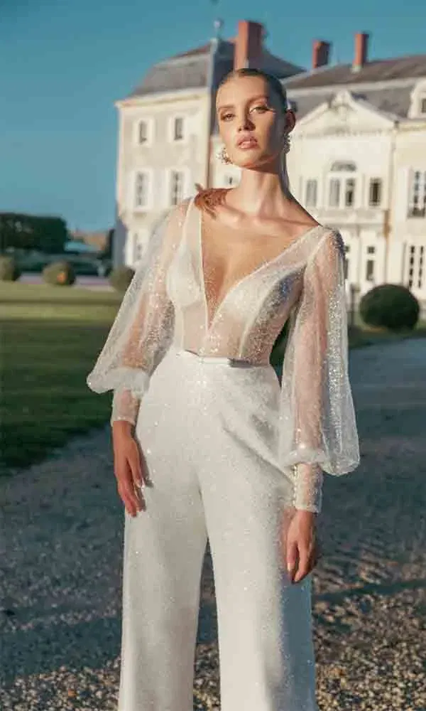 Nahaufnahme einer Braut im funkelnden Jumpsuit mit zartem Tüll und Schleifendetail, aufgenommen im Abendlicht vor einem Schloss.
