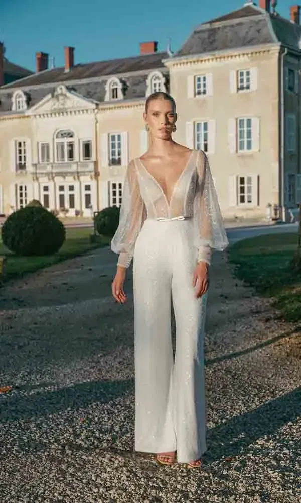 Glänzender Hochzeitslook mit weiter Hose und tiefem Ausschnitt – die Braut posiert vor einem eleganten Landsitz bei Tageslicht.