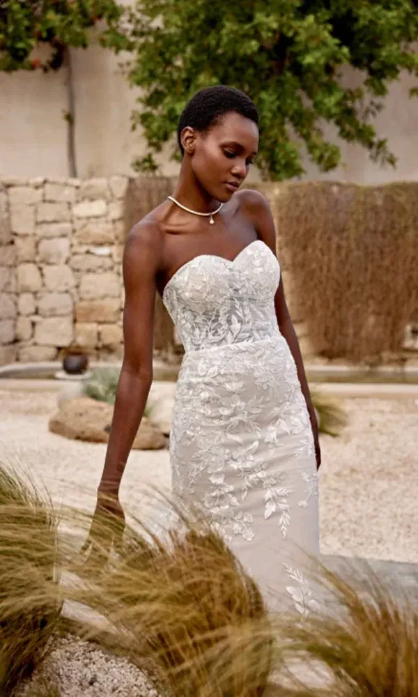 Stilvolles Porträt einer Braut mit Herzausschnitt in sommerlicher Umgebung mit Kiesboden und Steinmauer im Hintergrund.