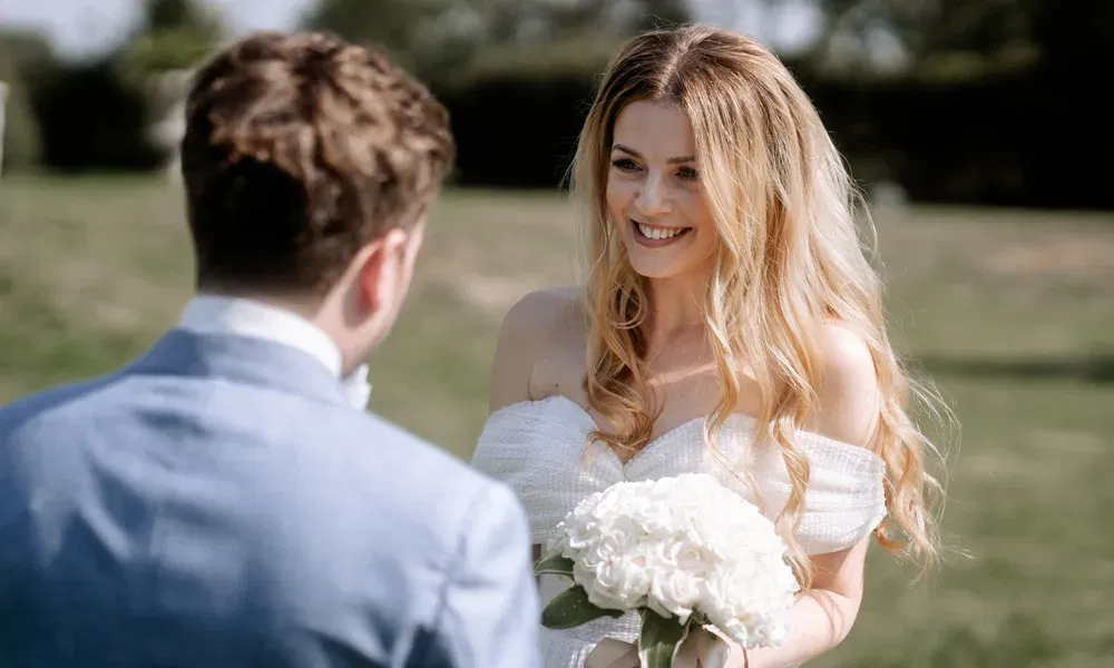 Auf einer sonnigen Wiese lächelt die Braut in einem schulterfreien Kleid ihren Bräutigam an und hält dabei einen Strauß weißer Hortensien.
