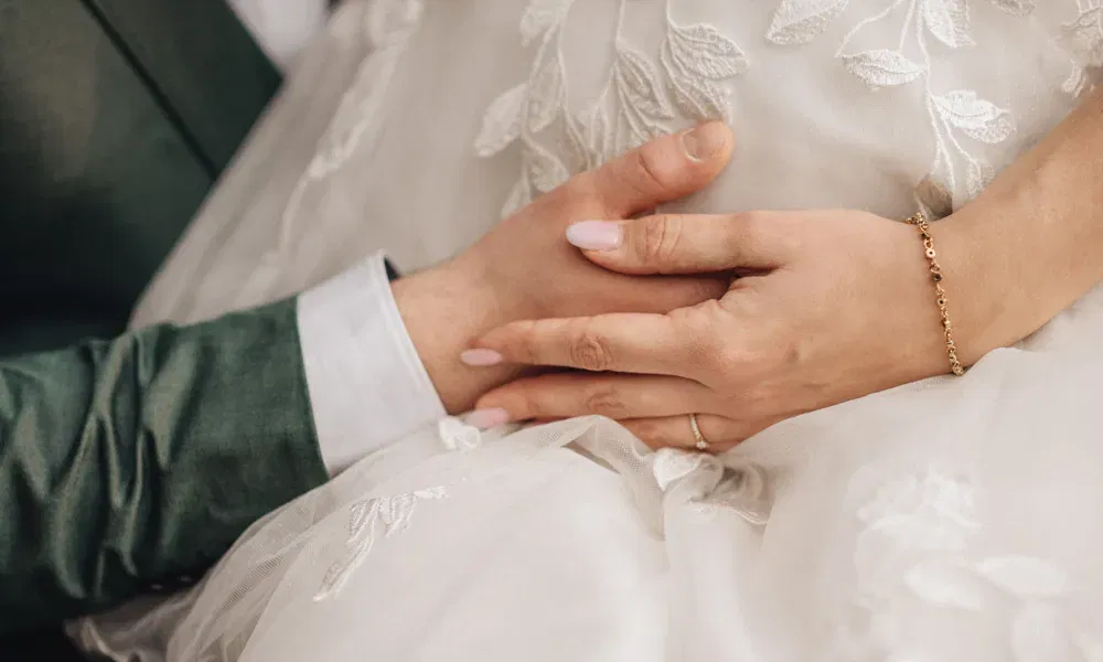 Hochzeitsszene in Nahaufnahme: Bräutigam und Braut legen schützend die Hände auf ihren Babybauch, umgeben von zarter Spitze.