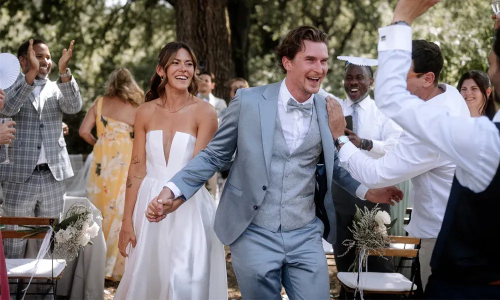 Ausgelassene Szene nach der Zeremonie: Das Brautpaar feiert im Freien unter Bäumen, umgeben von gratulierenden Hochzeitsgästen.