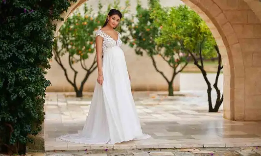 Frau im Empire-Brautkleid mit Cap-Ärmeln und Spitze posiert vor blühenden Bäumen in einem steinernen Arkadengang.