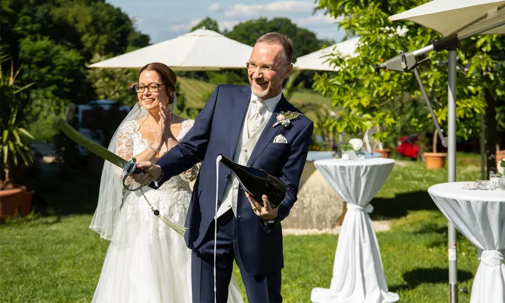 Bräutigam öffnet mit einem Säbel eine Champagnerflasche, während die Braut lachend zuschaut.