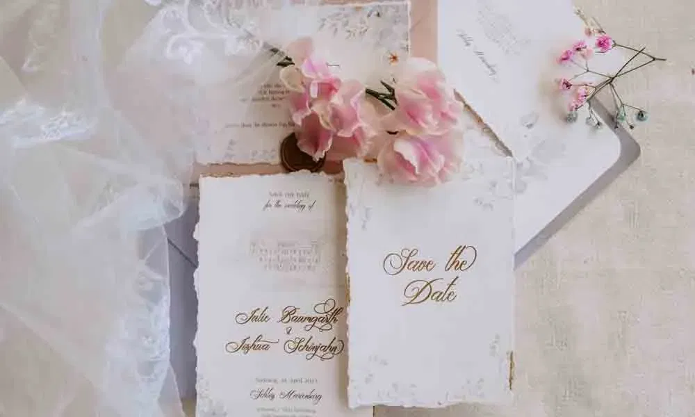 Verspieltes Flatlay mit Save-the-Date-Karten, rosa Blüten und Spitzenstoff im Stil einer Bridgerton-Hochzeit.