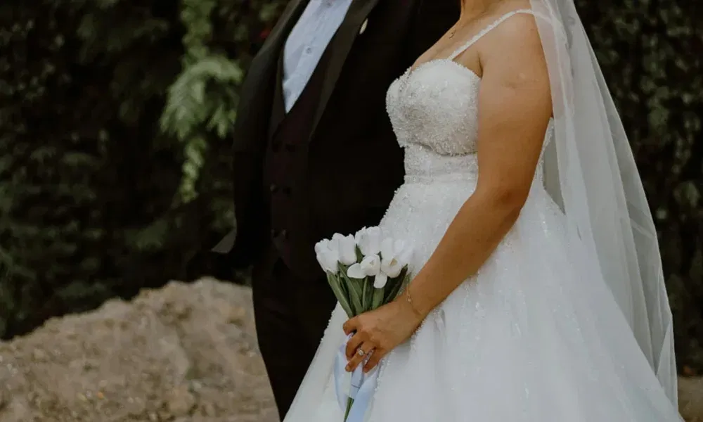 Nahaufnahme der Braut in einem schimmernden weißen Kleid mit Spaghettiträgern und Brautstrauß, der Bräutigam teilweise sichtbar.