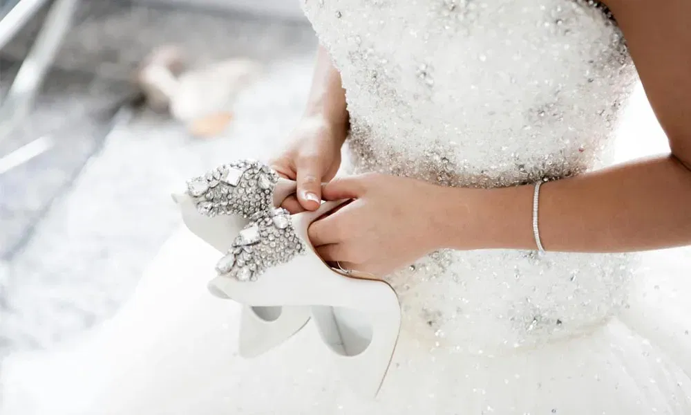 Braut im festlichen Hochzeitskleid bereitet sich vor und trägt Schmuck, während sie funkelnde, mit Kristallen besetzte Schuhe in Händen hält.