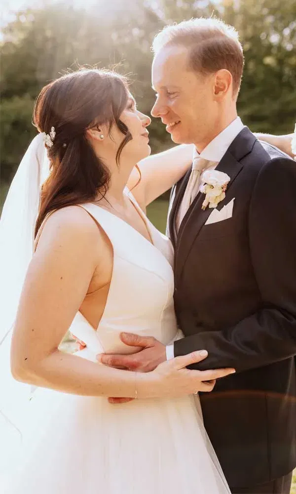 Braut in weißem Kleid und Bräutigam im Anzug halten sich fest und sehen sich voller Liebe an, die Sonne scheint durch die Bäume.