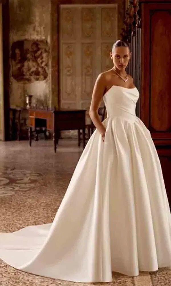 Zeitlose Brautmode: Frau in luxuriösem, trägerlosen Brautkleid mit Faltenrock und Schleppe in stilvoller Altbaukulisse.