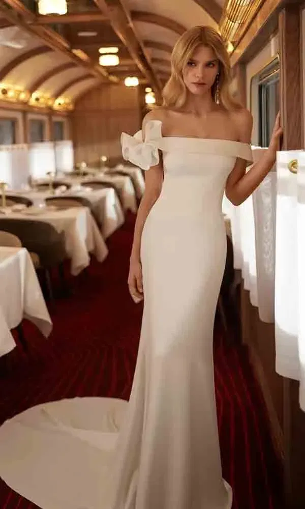 Modernes Hochzeitskleid mit schulterfreier Passform und dekorativer Schleife, aufgenommen in einem luxuriösen Waggon mit rotem Teppich.