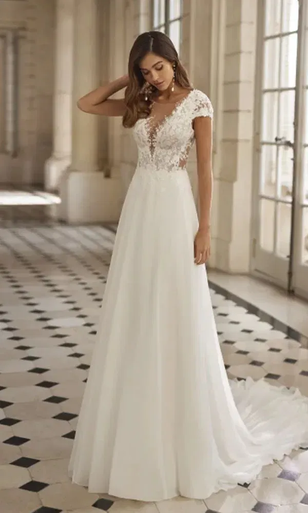 Romantischer Brautlook in einem luftigen A-Linien-Kleid mit Spitzenoberteil und Cap-Ärmeln, eingefangen in lichtdurchfluteter Kulisse.